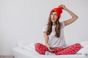 garota engraçada sorridente em um chapéu vermelho e pijama de natal, sentado na cama com lençóis brancos. feriados para o natal. garota de bom humor natalino. menina em uma camiseta branca e pijama, foto