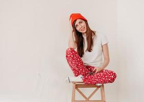 garota engraçada sorridente em um chapéu vermelho e pijama de natal, sentado na cama com lençóis brancos. feriados para o natal. garota de bom humor natalino. garota em uma camiseta branca e pijama foto