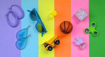 composição pastel na moda com brincos, óculos de sol, lata de bebida, bola de basquete, caminhão de brinquedo, caixa de presente, flor e girador nas cores rosa, violeta, verde, laranja, amarelo e azul foto
