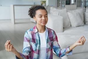 garota calma de raça mista medita para aliviar o estresse, faz exercícios respiratórios com gesto de mudra em casa foto