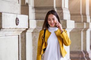 retrato de uma mulher asiática jovem, atraente e bonita sorrindo enquanto ela fala em seu smartphone na cidade. ela está desviando o olhar enquanto fala animada e alegremente em seu dispositivo móvel.