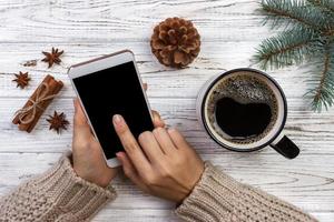 garota com um suéter de malha aconchegante usa o telefone enquanto está sentado em uma mesa com acessórios de natal foto