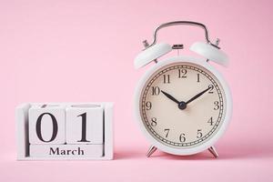 despertador e blocos de calendário de madeira com data 1 de março no fundo rosa.