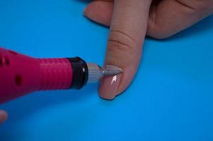 o processo de fazer uma bela manicure nos dedos de um dedo processando uma unha com uma máquina especial em um salão de beleza de unhas em um fundo azul foto