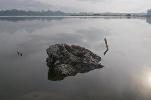 vista frontal de uma grande rocha no lago karangkates indonésia com condições de águas calmas ao nascer do sol com um fundo de montanhas e um céu nublado foto