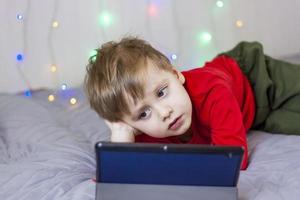 um menino bonito está na cama olhando para um tablet. treinamento online, comunicação na internet. criança sorridente engraçada. foto