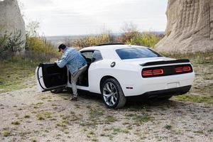 homem bonito em jaqueta jeans e boné sentado em seu muscle car branco na carreira. foto