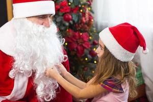 menina de chapéu de papai noel no colo do papai noel perto da árvore de natal na decoração de natal. mostra uma caixa com um presente, um peluche, mexe na barba e ri. ano novo, geada do avô russo foto