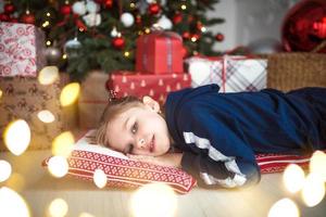 uma garotinha com roupas de casa está deitada debaixo da árvore de natal perto das caixas com presentes no travesseiro e sonhos. luzes de guirlandas, véspera de natal, ano novo. foto