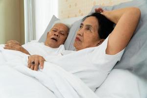mulher asiática mais velha que abre os olhos na cama ao lado do marido que ronca e faz barulho, problemas conjugais. foto