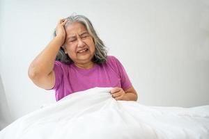 idosa insalubre mulher asiática sênior sentada na cama sofrendo de dor de cabeça e enxaqueca, então dor e doença, estresse mulher idosa sentada na cama em casa sozinha, experimentada, conceito de desânimo de idosos infelizes foto