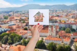 a mão de uma mulher está segurando um papel com uma casa recortada, panorama da cidade ao fundo. o conceito de alugar ou comprar imóveis foto