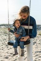 jovem mãe segurando seu filho sentado no balanço na praia. foto