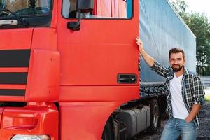 jovem motorista de caminhão sorridente ao lado de seu caminhão de carga vermelho foto
