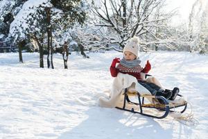 menino da criança sentado no trenó em um parque da cidade de neve durante o dia ensolarado de inverno foto