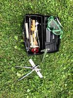 uma caixa de ferramentas preta fica no chão, em um gramado verde. dentro da caixa, várias ferramentas são armazenadas para reparos e para a construção de uma casa. alicate, um nível, óculos para cortar grama estão por perto foto