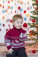 uma criança está segurando um brinquedo de árvore de natal enquanto está sentada sob uma elegante árvore de natal. emoções das crianças. natal e ano novo. foto