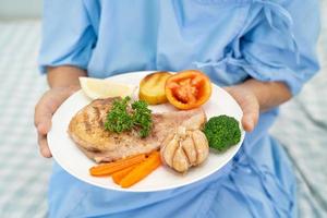 paciente idosa asiática ou idosa tomando café da manhã e comida saudável vegetal com esperança e feliz enquanto está sentado e com fome na cama no hospital. foto