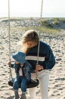 jovem mãe segurando seu filho sentado no balanço na praia. foto