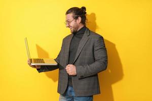 homem barbudo feliz usando óculos usando computador portátil em fundo amarelo foto