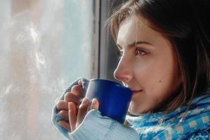 mulher bonita e triste ficando em casa por causa da gripe e temperatura no suéter quente e cachecol com xícara de chá perto da janela foto