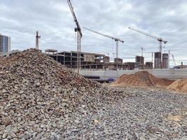 construção de casas altas na cidade. guindastes puxam lajes de concreto pesadas para a construção de edifícios residenciais. pedras para estradas são derramadas ao redor foto