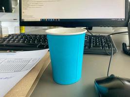 xícara de café azul de papelão de uma cafeteria em uma área de trabalho com um computador com teclado e mouse e material de escritório de um trabalhador de escritório durante o almoço. pausa para café de negócios foto