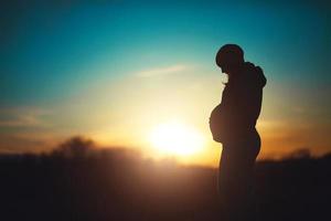 silhueta de uma futura mãe, mulher grávida relaxa no parque em um fundo por do sol foto