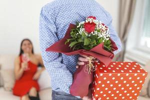 momento inesperado na rotina da vida cotidiana foto recortada das mãos do homem se escondendo segurando buquê chique de rosas vermelhas e caixa de presente nas costas, mulher feliz está no fundo desfocado