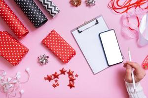 folha de papel em branco, telefone e mãos femininas embrulhando presentes de natal deitados sobre fundo rosa foto