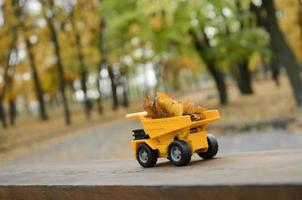 um pequeno caminhão amarelo de brinquedo é carregado com folhas amarelas caídas. o carro fica em uma superfície de madeira contra um fundo de um parque de outono embaçado. limpeza e remoção de folhas caídas. trabalhos sazonais foto