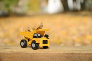 um pequeno caminhão amarelo de brinquedo é carregado com folhas amarelas caídas. o carro fica em uma superfície de madeira contra um fundo de um parque de outono embaçado. limpeza e remoção de folhas caídas. trabalhos sazonais foto