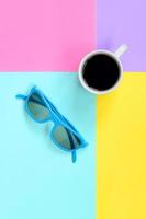 pequena xícara de café branco e óculos de sol azuis sobre fundo de textura de papel de cores azul, amarelo, violeta e rosa pastel de moda em conceito mínimo foto