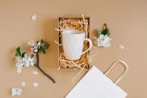 uma caixa de presente aberta com aparas de papel cor de pêssego contendo uma caneca criativa da moda com um padrão geométrico, um saco de papel branco e flores de maçã em um fundo bege. latitude plana, vista superior foto