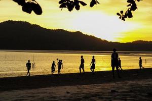 grupo de silhueta de pessoas jogando futebol na praia por do sol com vista do mar e montanha ao fundo foto