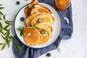 panquecas fofas tradicionais com geléia de laranja, mirtilos, lascas de coco e hortelã para café da manhã saudável, luz de fundo. fotografia chave alta. foto