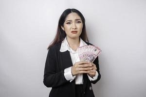 uma jovem empresária confusa está vestindo terno preto e segurando dinheiro em rúpia indonésia isolada por fundo branco foto