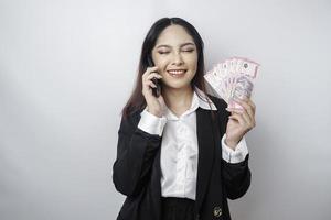 uma jovem empresária feliz está vestindo terno preto, segurando seu telefone e dinheiro em rupia indonésia isolado por fundo branco foto