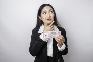 uma jovem pensativa está vestindo terno preto e segurando dinheiro em rúpia indonésia isolada por fundo branco foto