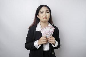 uma jovem empresária confusa está vestindo terno preto e segurando dinheiro em rúpia indonésia isolada por fundo branco foto