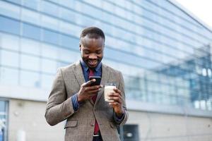 homem afro-americano feliz lendo algo em seu smartphone enquanto está do lado de fora com uma xícara de café