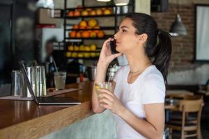 jovem mulher encantadora ligando com o telefone celular enquanto está sentada sozinha na cafeteria durante o tempo livre, mulher atraente com sorriso fofo conversando com o celular enquanto descansa no bar foto