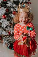 uma garotinha de cabelos cacheados em um vestido de carnaval escondeu o rosto atrás de estrelas brilhantes de brinquedos de árvore de natal. criança em um vestido vermelho com uma estampa de papai noel no fundo de uma árvore de natal. foto de alta qualidade