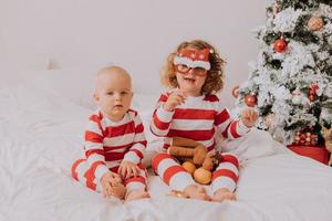 crianças de pijama vermelho e branco experimentam óculos engraçados com papai noel sentado na cama. estilo de vida. irmão e irmã comemorando o natal. menino e menina estão brincando em casa. foto de alta qualidade