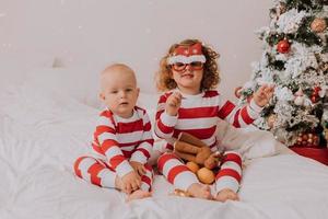 crianças de pijama vermelho e branco experimentam óculos engraçados com papai noel sentado na cama. estilo de vida. irmão e irmã comemorando o natal. menino e menina estão brincando em casa. foto de alta qualidade