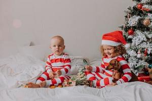 crianças de pijama vermelho e branco sentadas na cama compartilham doces de natal umas com as outras e com o cachorro. irmão e irmã, menino e menina comemoram o ano novo. espaço para texto. foto de alta qualidade
