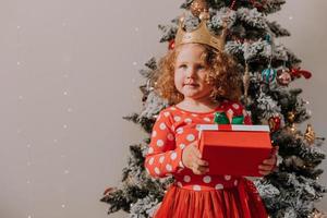 uma garotinha de cabelos cacheados em um vestido de carnaval escondeu o rosto atrás de estrelas brilhantes de brinquedos de árvore de natal. criança em um vestido vermelho com uma estampa de papai noel no fundo de uma árvore de natal. foto de alta qualidade