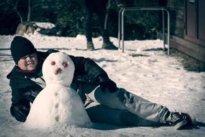 menino deitado na neve ao lado de um boneco de neve feito por ele mesmo com nariz de cenoura e olhos de tomate foto