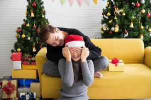 homem bonito preparando uma surpresa de presente de natal para linda mulher de chapéu de papai noel com os olhos fechados, conceito de cartão-presente foto