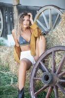 garota sensual com pernas longas e botas marrons senta na cadeira de madeira na fazenda. garota bonita vestindo corpo bonito na cena ocidental ao ar livre. moda modelo loira com pernas longas, sutiã e cabelos cacheados foto